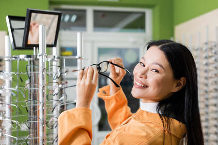 asian woman smiling at camera while holding eyeglasses near mirror in optics shopの写真素材