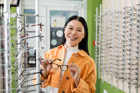 cheerful asian woman holding eyeglasses while looking at camera in optics shop
