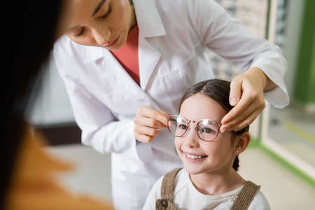 ophthalmologist trying eyeglasses on smiling girl near blurred mom in optics store