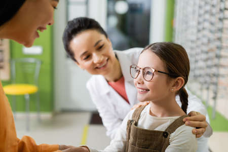 happy girl in eyeglasses smiling near asian mother and blurred oculist in optics storeの写真素材