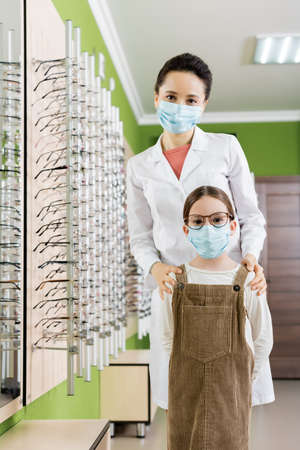 oculist and girl in medical masks looking at camera near assortment of eyeglasses in optics store