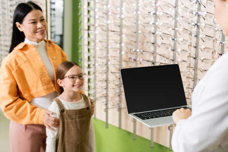 blurred interracial mother and daughter smiling near oculist with laptop in optics storeの写真素材