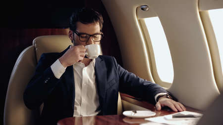 Businessman in eyeglasses drinking coffee near smartphone and papers in private planeの写真素材