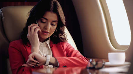 Businesswoman talking on smartphone and looking at wristwatch near cup in private plane