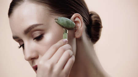 young woman using jade roller isolated on beigeの写真素材