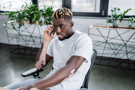 young african american man with vitiligo and trendy hairstyle talking on smartphone in office