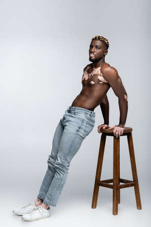 shirtless african american man with vitiligo and muscular torso posing in jeans near high stool on greyの写真素材