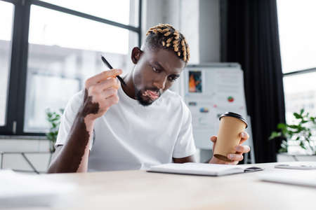 serious african american man with vitiligo holding paper cup and pen while looking in notebook in officeの写真素材
