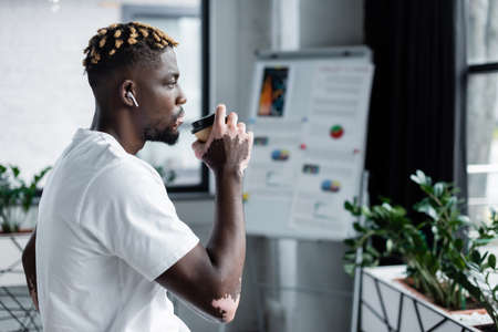 side view of african american man with vitiligo drinking coffee and listening music in earphone near...