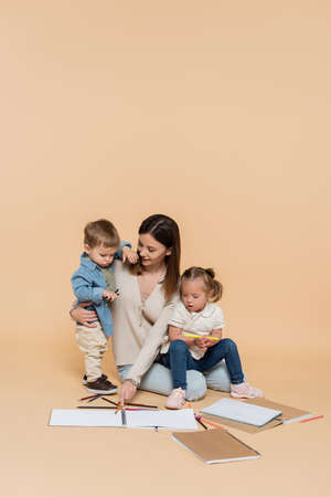 cheerful mother sitting near girl with down syndrome, toddler boy and colorful pencils on beigeの写真素材