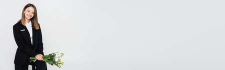 cheerful woman in suit holding bouquet of flowers and sitting on chair isolated on grey, bannerの写真素材