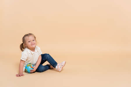 happy kid with down syndrome sitting with small globe on beigeの写真素材
