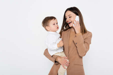 happy young woman in beige blazer talking on smartphone and holding in arms toddler boy isolated on greyの写真素材
