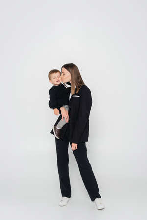 full length of mother holding in arms and kissing toddler son on greyの写真素材