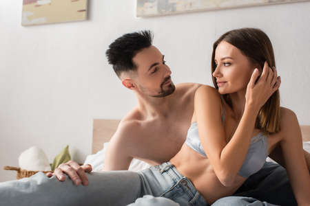 seductive woman in bra and jeans looking at shirtless man in bedroom