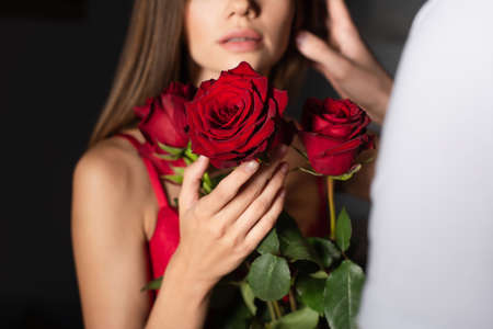 cropped view of blurred woman holding red roses near man on dark background