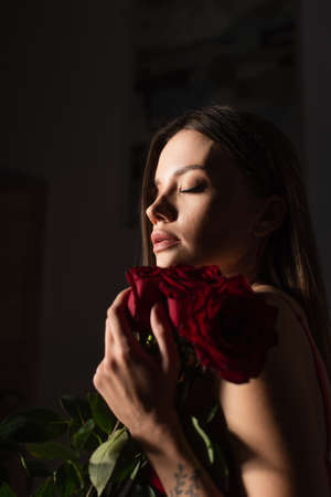 young and sensual woman with closed eyes holding red roses on dark backgroundの写真素材