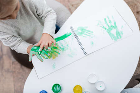 Top view of kid applying paint on hand near sketchbook on coffee tableの写真素材