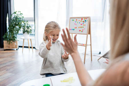 Blurred woman near smiling kid with paint on hands at home