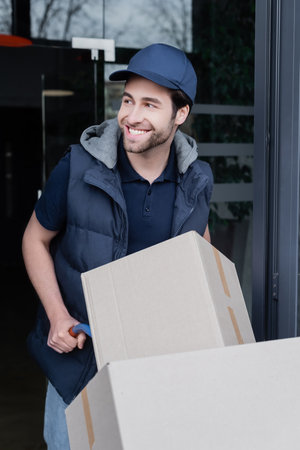 Smiling delivery man looking away near car with cardboard boxes and building outdoors