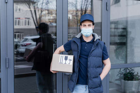 Young delivery man in medical mask holding cardboard box and looking at camera near building...