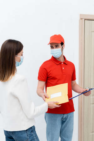 Courier in medical mask holding parcel and clipboard near customer in hallway
