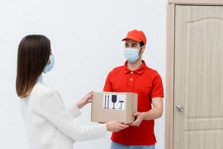 Delivery man in medical mask holding cardboard box near woman and door in hallwayの写真素材