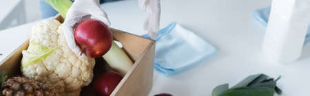 Cropped view of woman in latex gloves holding apple near food, rags and antiseptic at home, banner