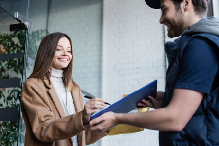 Smiling woman with parcel writing on clipboard near blurred courier in hallway