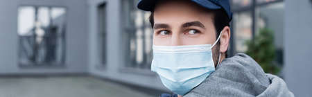 Courier in medical mask and cap looking away on urban street, banner