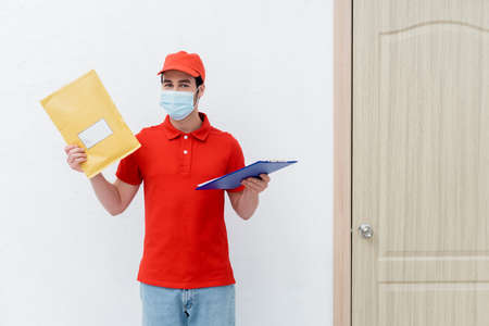 Courier in medical mask holding parcel and clipboard near door in hallway