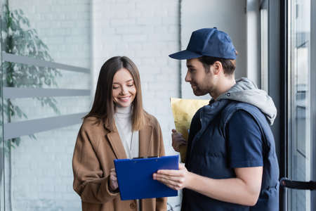Smiling woman looking at clipboard near courier with parcel in hallwayの写真素材