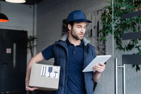 Delivery man in uniform holding cardboard box and using digital tablet in hallwayの写真素材