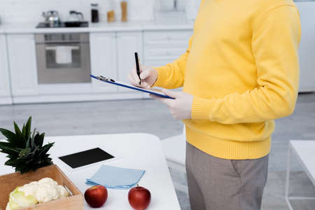 Cropped view of man in latex gloves writing on clipboard near fresh food and digital tablet in...