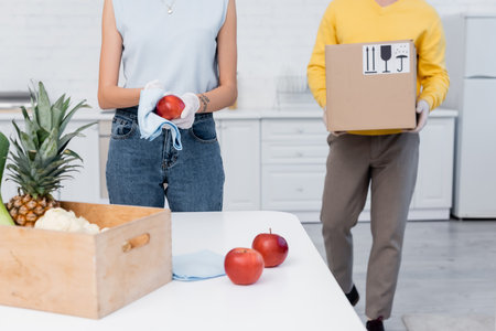 Cropped view of woman in latex gloves cleaning apple near boyfriend carrying cardboard box in...