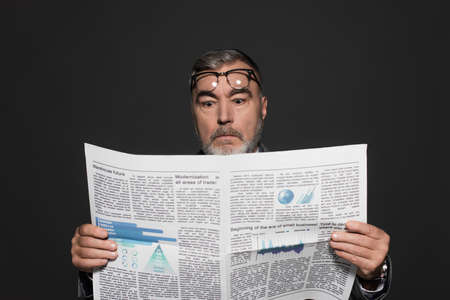 surprised senior businessman with eyeglasses on forehead reading newspaper isolated on dark grayの写真素材