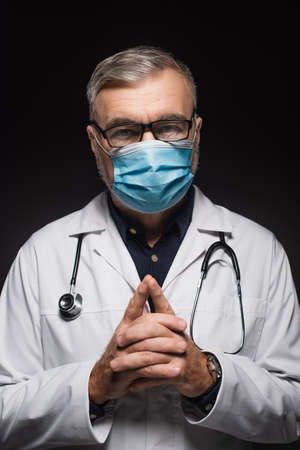 doctor in medical mask and eyeglasses standing with clasped hands and looking at camera isolated on...
