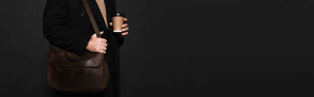 partial view of man in coat standing with paper cup and leather bag isolated on black, bannerの写真素材