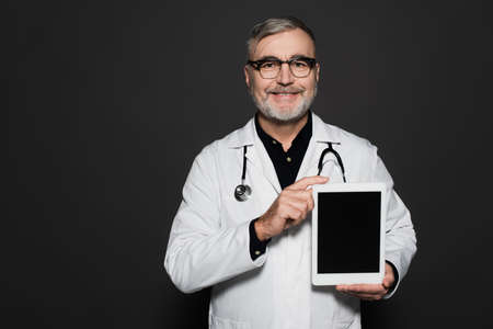 joyful senior doctor in white coat and eyeglasses holding digital tablet with blank screen isolated...
