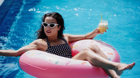 brunette woman in sunglasses holding glass with cocktail and swimming on inflatable ring in pool