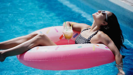 pleased woman in sunglasses holding glass with cocktail and swimming on inflatable ring in pool