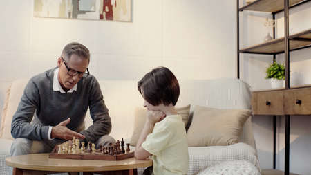grandfather in glasses and smart grandson playing chess in living roomの写真素材