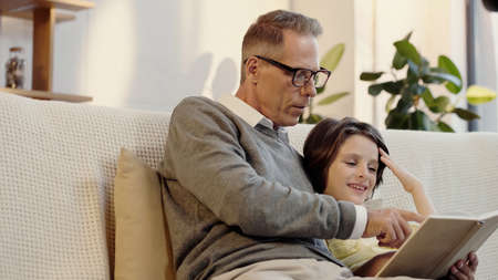 middle aged grandfather in glasses reading book with happy grandson at home