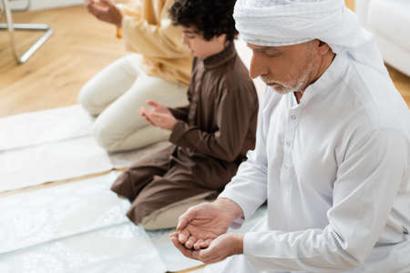 Mature man praying near blurred muslim grandson and son at home