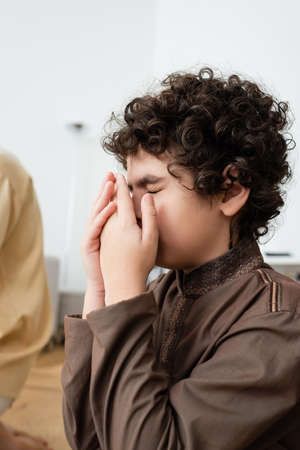 Curly arabian boy holding hands near face while praying near blurred father at homeの写真素材