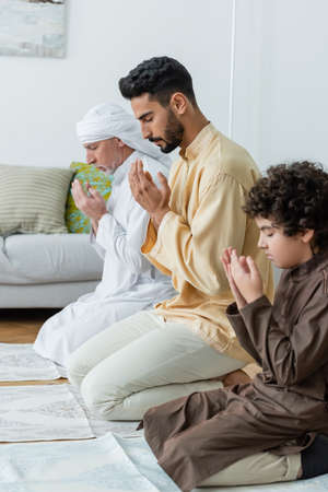 Side view of young muslim man praying near father and son on rugs at homeの写真素材