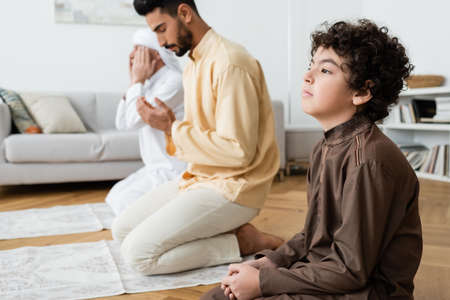 Curly muslim boy praying near blurred father and granddad on rugs at home
