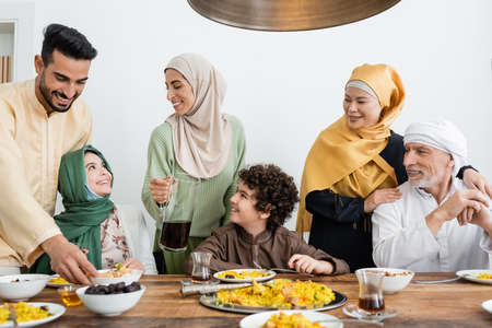 young arabian woman holding jug of tea near multiethnic muslim family having dinner at homeの写真素材
