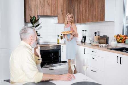 Smiling woman holding pie near blurred husband with champagne at homeの写真素材
