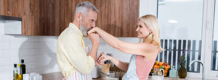 Middle aged man in apron kissing hand of smiling wife in kitchen, bannerの写真素材